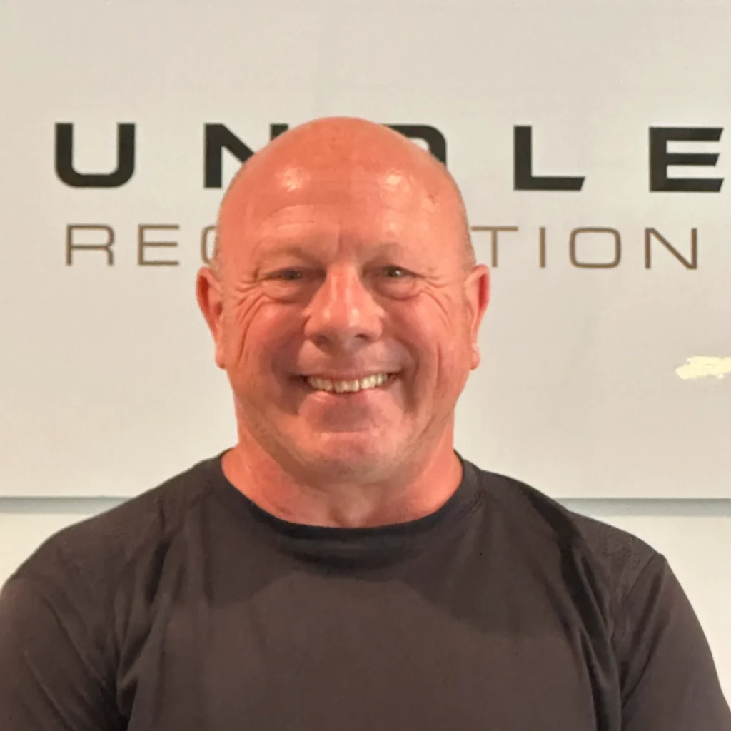 Smiling bald man in a black t-shirt standing in front of a wall with large letters behind him (partial logo)