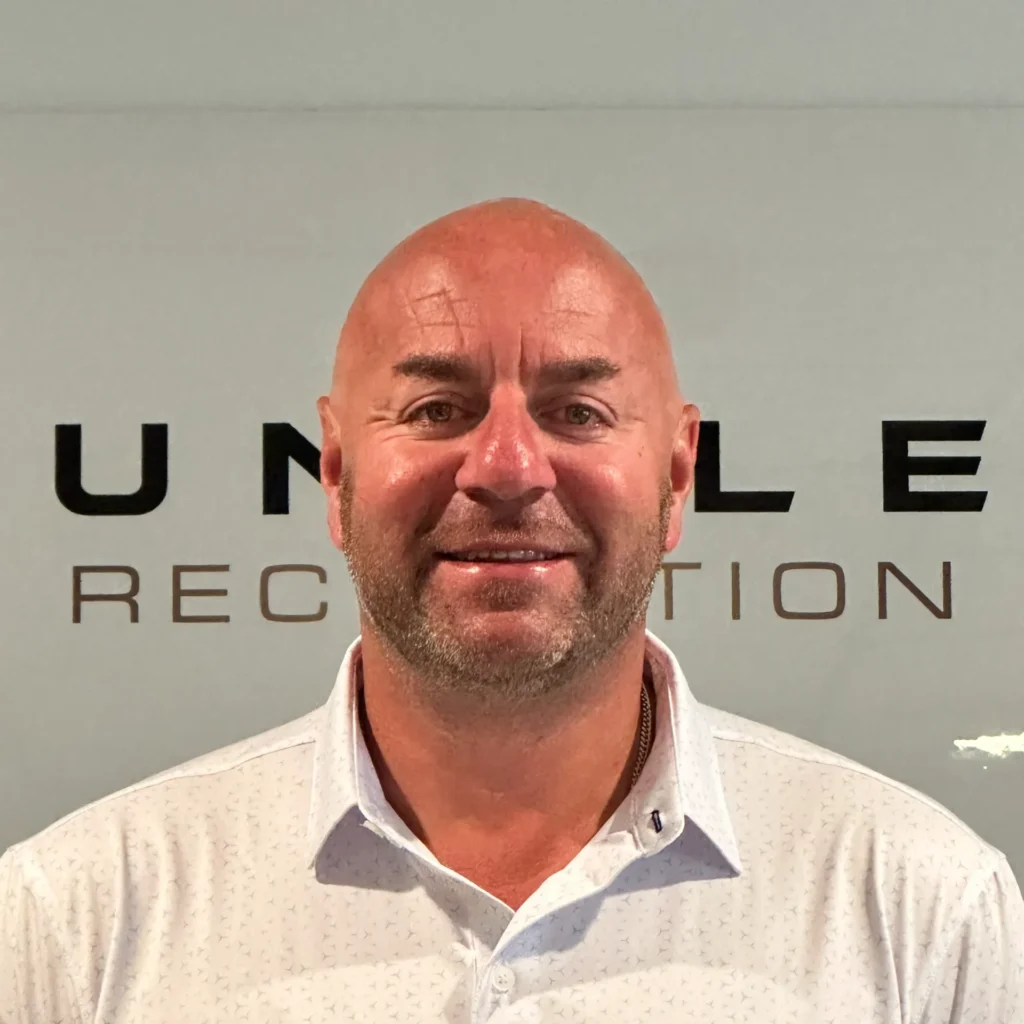 Head-and-shoulders mugshot of a bald man with stubble, posing against a gray backdrop with large black letters behind him.