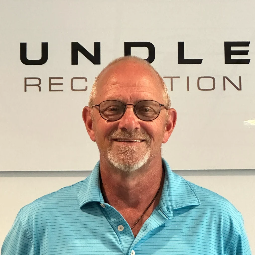 Smiling older man with glasses, wearing a teal striped polo, in front of a wall with large black letters behind him.