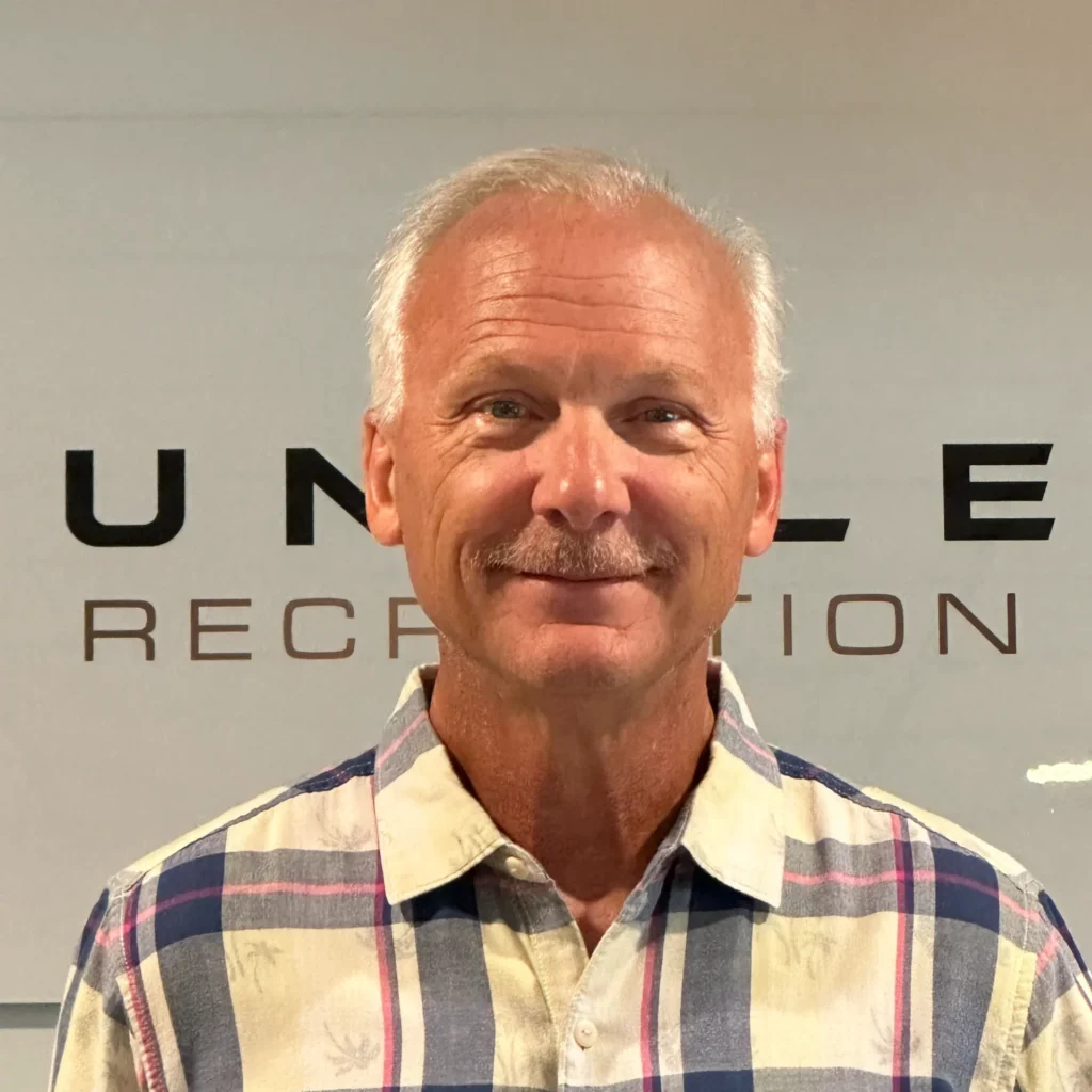 Partial head-and-shoulders portrait of a smiling older man with gray hair and a mustache, wearing a cream and blue plaid shirt, in front of a light wall with large black letters behind him