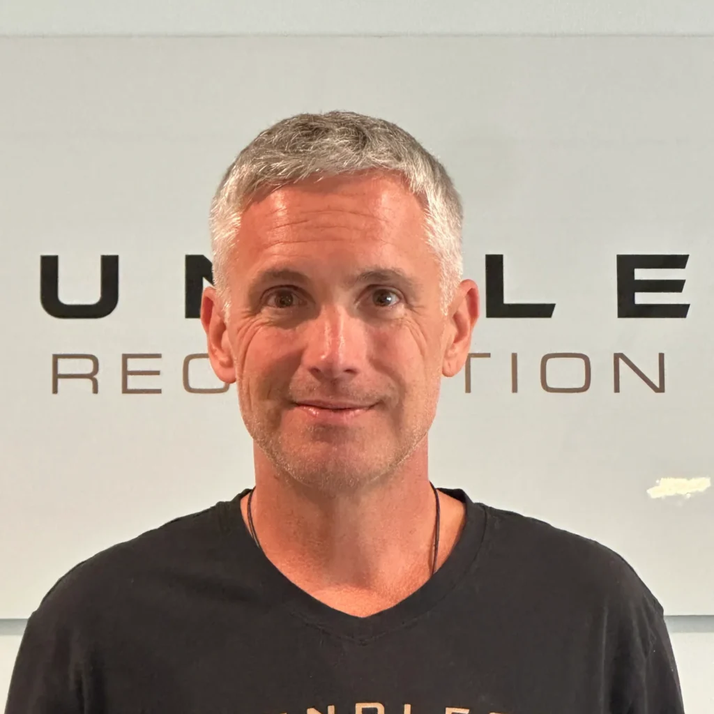 Close-up portrait of a smiling man with gray hair wearing a black shirt, standing in front of a light wall with large black text in the background.
