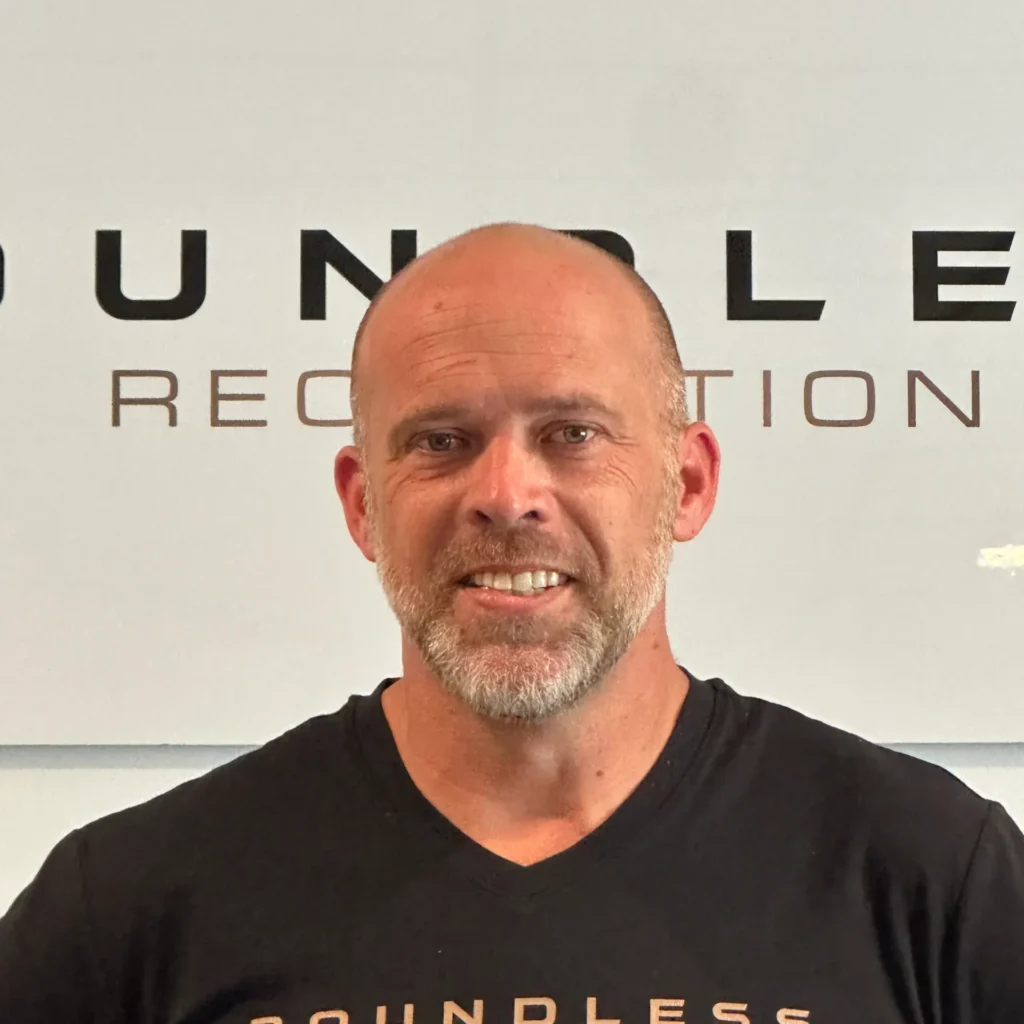 Smiling man taking a selfie in front of a light wall with large black letters behind him.