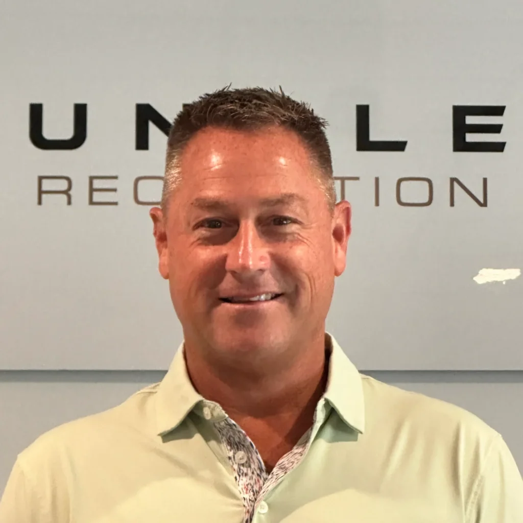 Smiling man in a beige shirt posing in front of a wall with large black letters behind him.